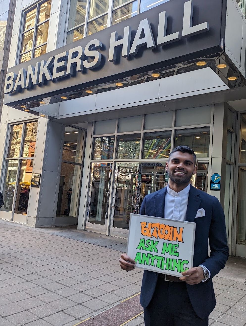 Santosh holding a Bitcoin, ask me anything sign outside Bankers Hall
