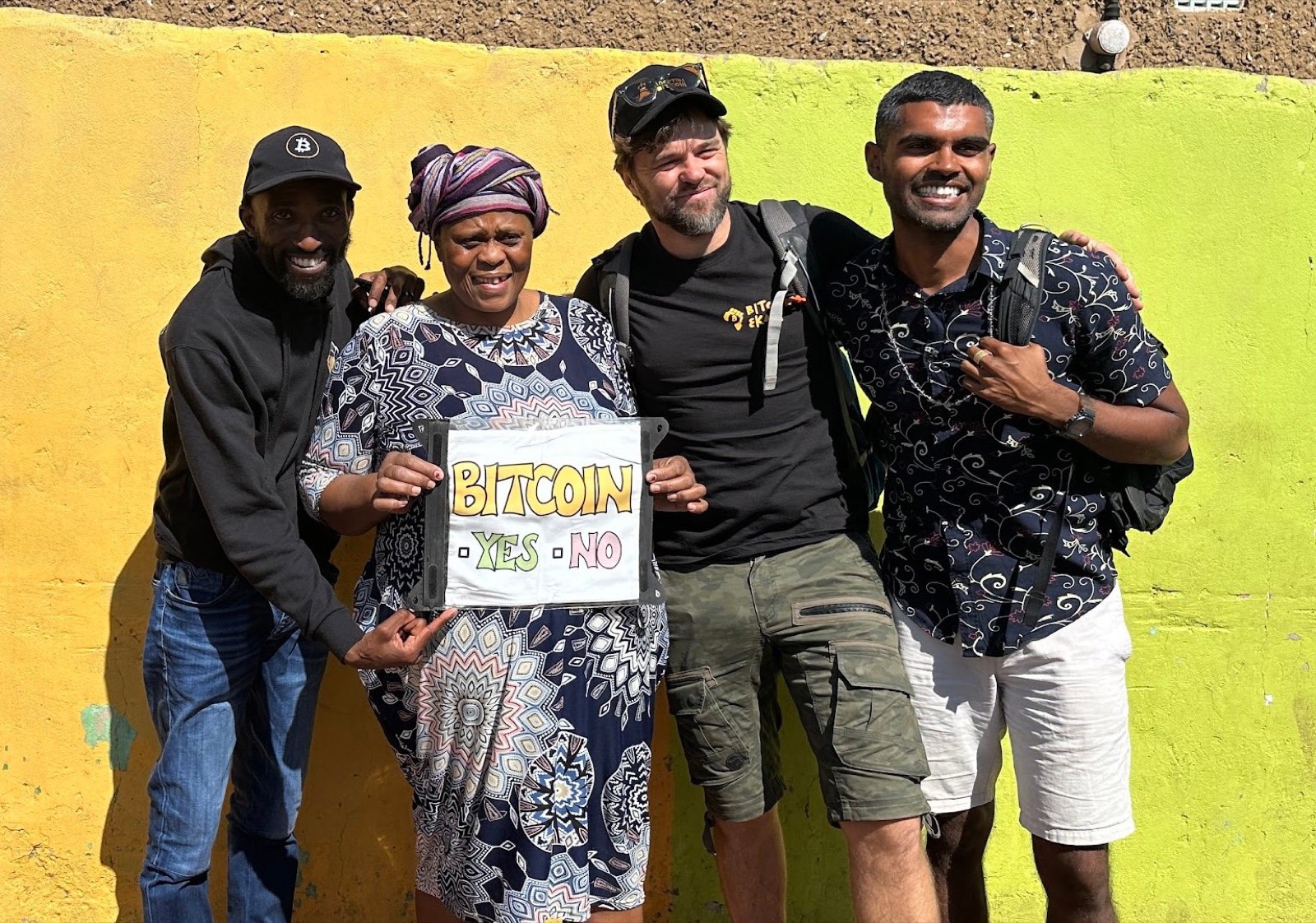 Four community members holding a hand-painted 'Bitcoin Yes No' sign against a yellow and green wall in Mohlakeng