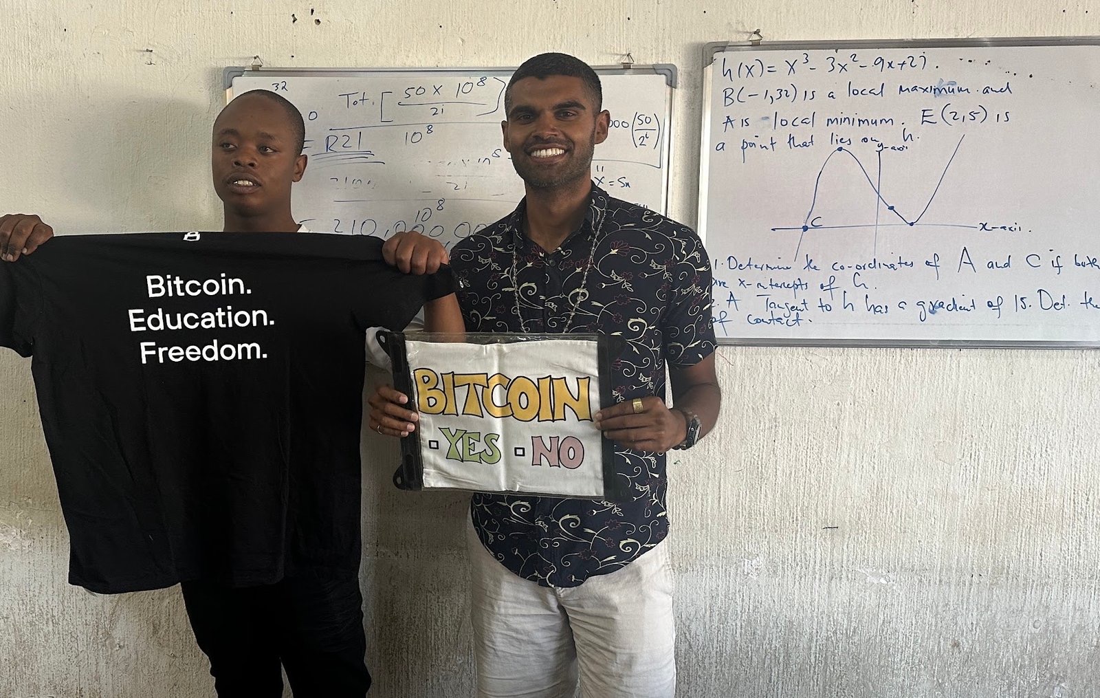 Two tutors at the math academy holding a 'Bitcoin. Education. Freedom.' t-shirt in front of a whiteboard of equations